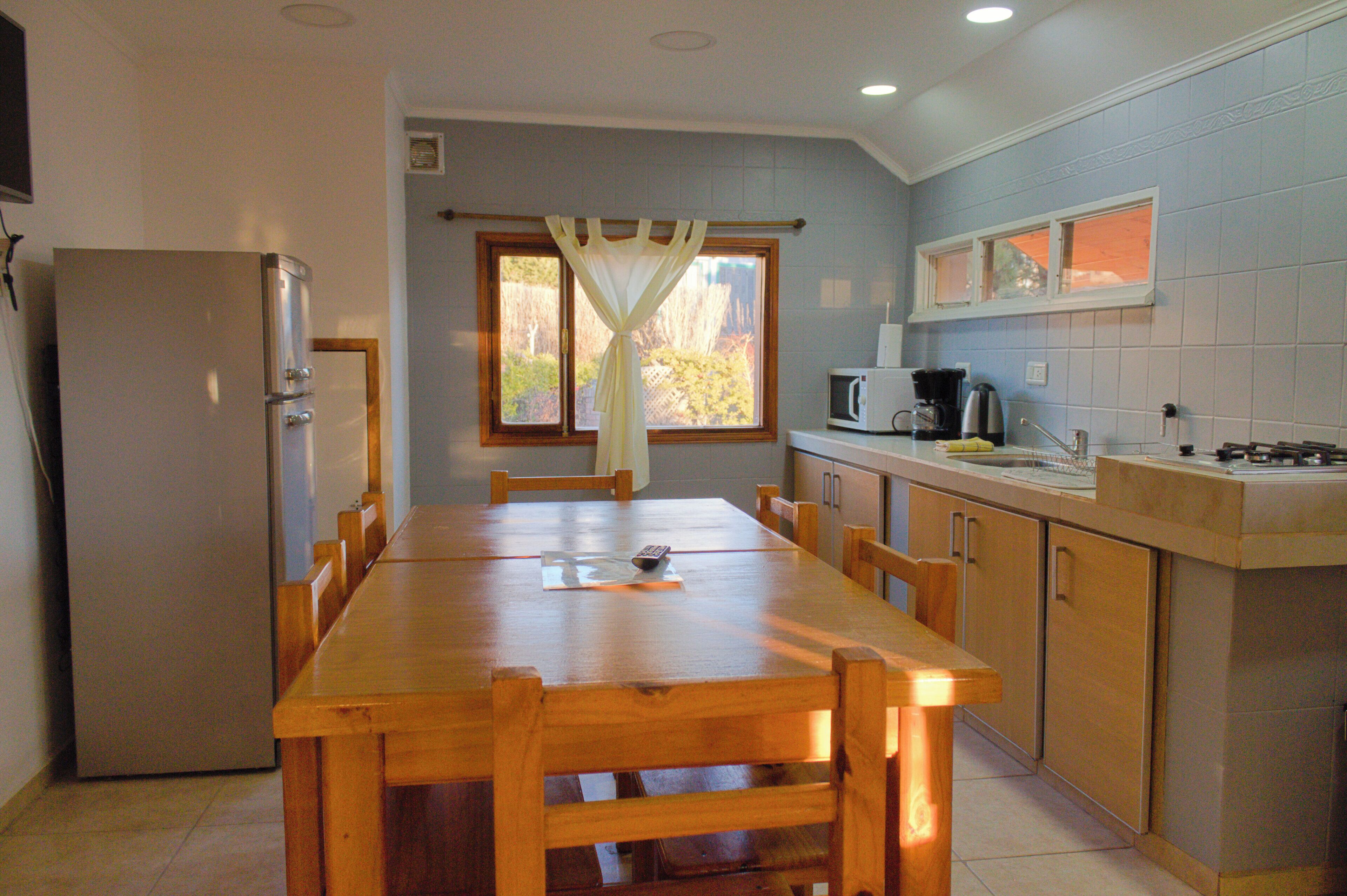 Una cocina con mesa de madera, armarios, un refrigerador y una ventana con cortinas.