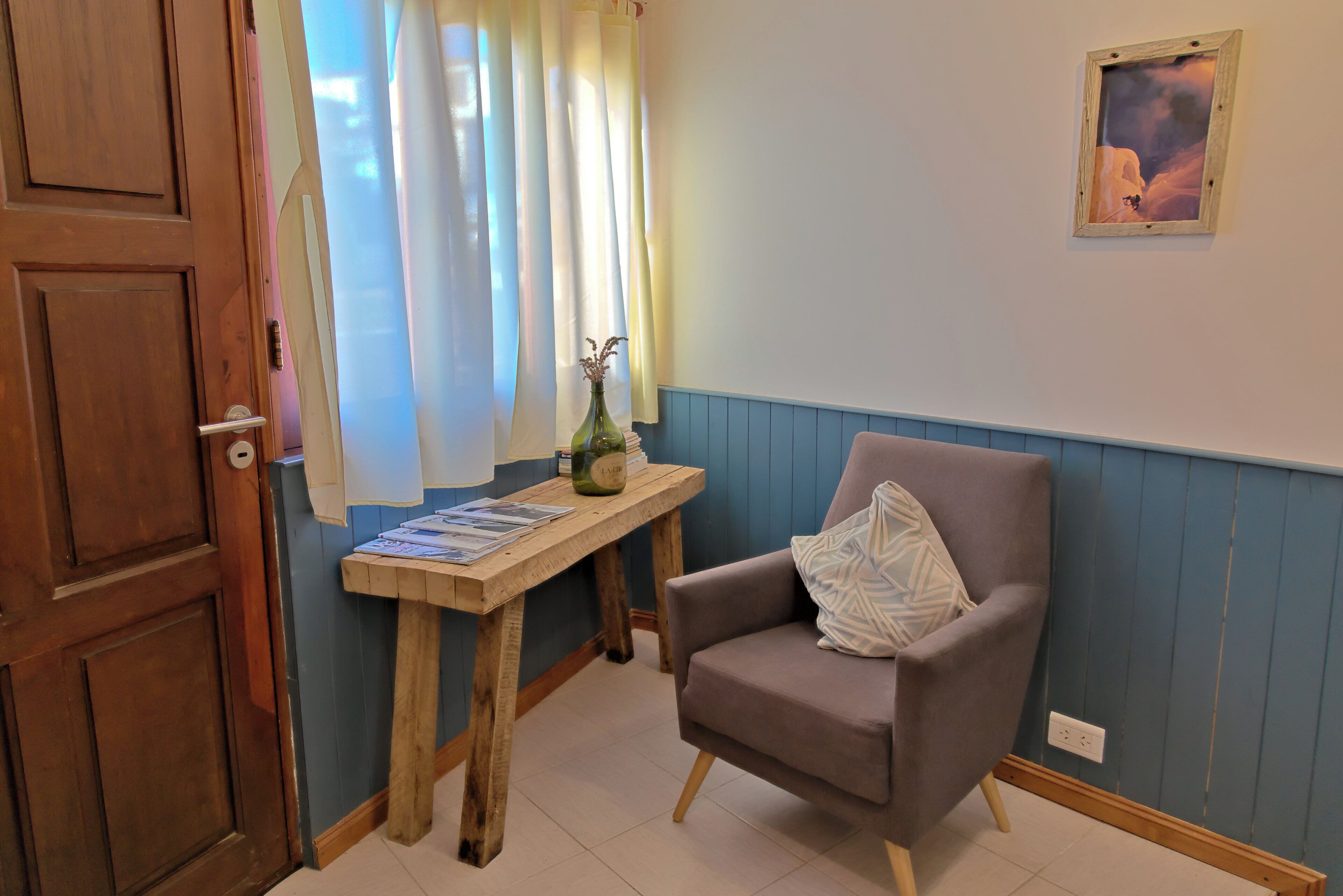 Una habitación con puerta de madera, una mesita con revistas, una silla con cojín y una ventana con cortinas traslúcidas.