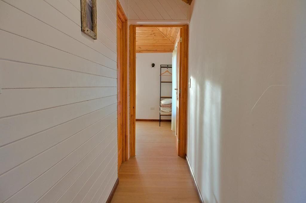 A narrow hallway with wooden flooring and white walls, leading to a room with a bed and a hanging rack.