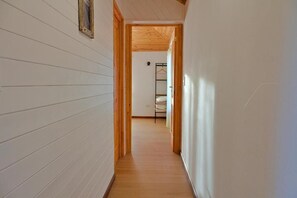 A narrow hallway with wooden flooring and white walls, leading to a room with a bed and a hanging rack.