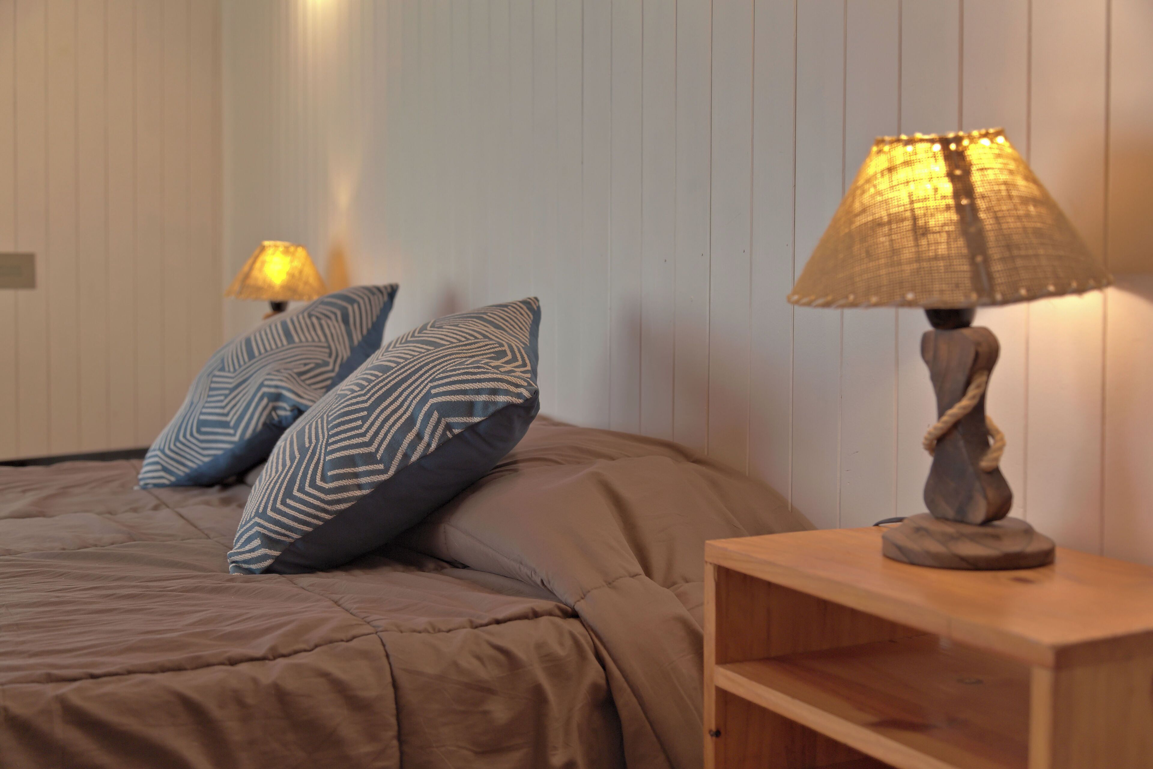 Un dormitorio con una cama, dos almohadas estampadas, una lámpara de noche y una mesita de noche de madera.