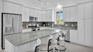A modern kitchen with white cabinetry, a central island with bar stools, stainless steel appliances, and a view of palm trees through a window. - Coco Plum Getaway - Dockage -short walk to Coco Plum Beach - Brand New Home! (Marathon)
