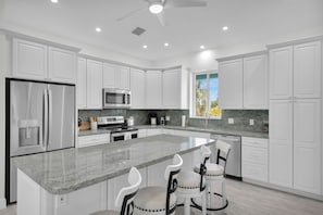 A modern kitchen with white cabinetry, a central island with bar stools, stainless steel appliances, and a view of palm trees through a window. - Coco Plum Getaway - Dockage -short walk to Coco Plum Beach - Brand New Home! (Marathon)