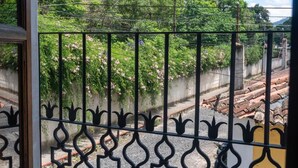Une vue à travers une grille en métal ornée d’un motif décoratif, donnant sur une rue bordée d’un mur en béton et surmontée d’un toit en tuiles.
