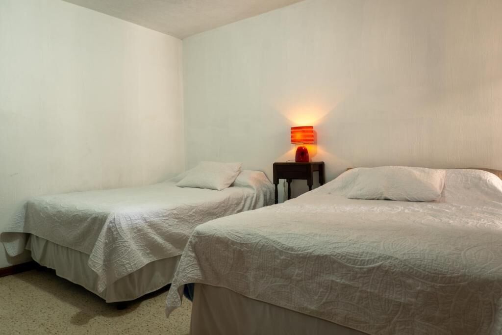 Quarto com duas camas de solteiro, mesa de cabeceira com abajur e parede branca lisa.