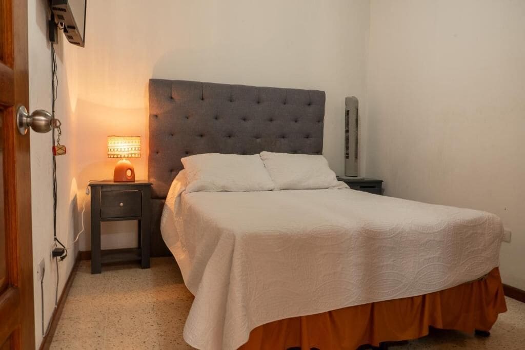 A bedroom with a bed, a nightstand, a lamp, and a wall-mounted heater.