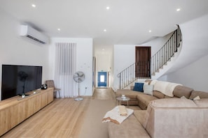 A modern living room with a flat-screen TV, a wooden entertainment unit, a beige sofa, a standing fan, and a staircase leading to an upper floor.