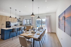 Une cuisine moderne dotée d’un îlot central, d’un coin repas avec des chaises, d’un éclairage suspendu et offrant une vue sur les bâtiments à travers de grandes fenêtres.