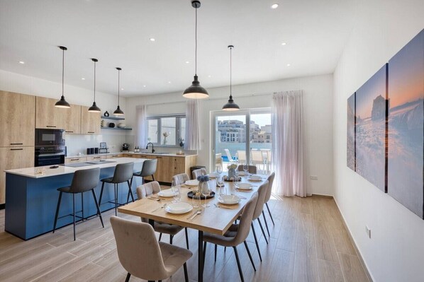 A modern kitchen with a central island, dining area with chairs, pendant lighting, and a view of buildings through large windows.