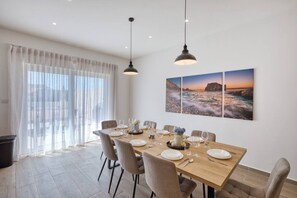 A dining area with a wooden table set for six, surrounded by chairs, with a large window and curtains, and two wall-mounted pendant lights.