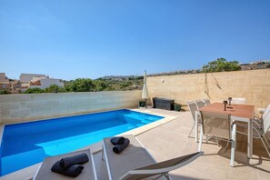 A rooftop swimming pool with a table and chairs, surrounded by a stone wall and greenery.