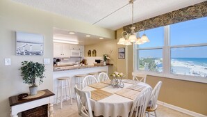 A dining area with a table set for four, white chairs, a chandelier, and a view of the beach through large windows. - Beachside Paradise - Views, Pool & Sunshine (New Smyrna Beach)