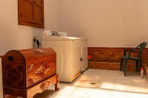 A laundry area with a washing machine, a wooden chest, and a plastic chair.