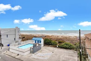 A beachfront property with a swimming pool, a small building with a blue roof, and a parking area. - Ocean View Oasis | Walk to Carolina Beach! (Carolina Beach)