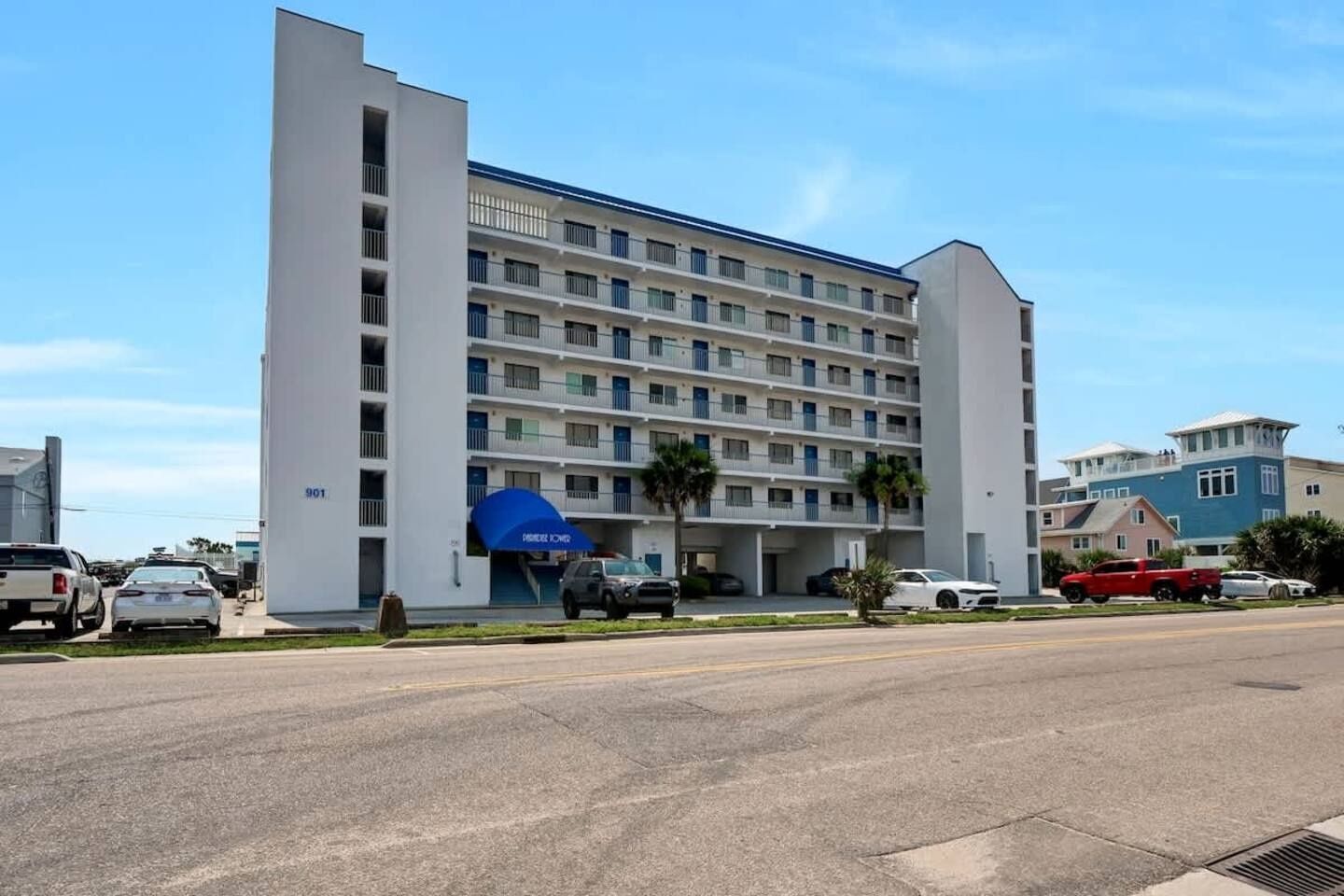 A multi-story hotel with a blue awning, parking lot, and a clear sky.