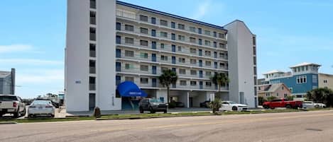 Hotel de vários andares com toldo azul, estacionamento e céu claro.