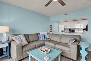 A living room with a sofa, a coffee table, a lamp, and a kitchen area in the background. - Ocean View Oasis | Walk to Carolina Beach! (Carolina Beach)
