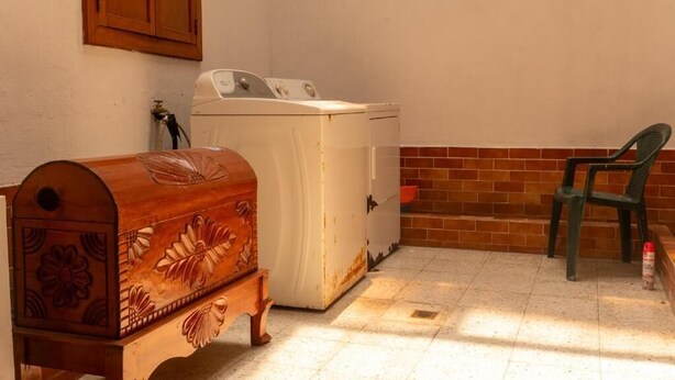 Un área de lavandería con lavadora, un cofre de madera y una silla de plástico.