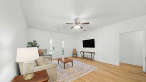 Ein großzügiges Wohnzimmer mit Deckenventilator, einer Couch, einem Couchtisch, einem an der Wand befestigten Fernseher und Holzboden.