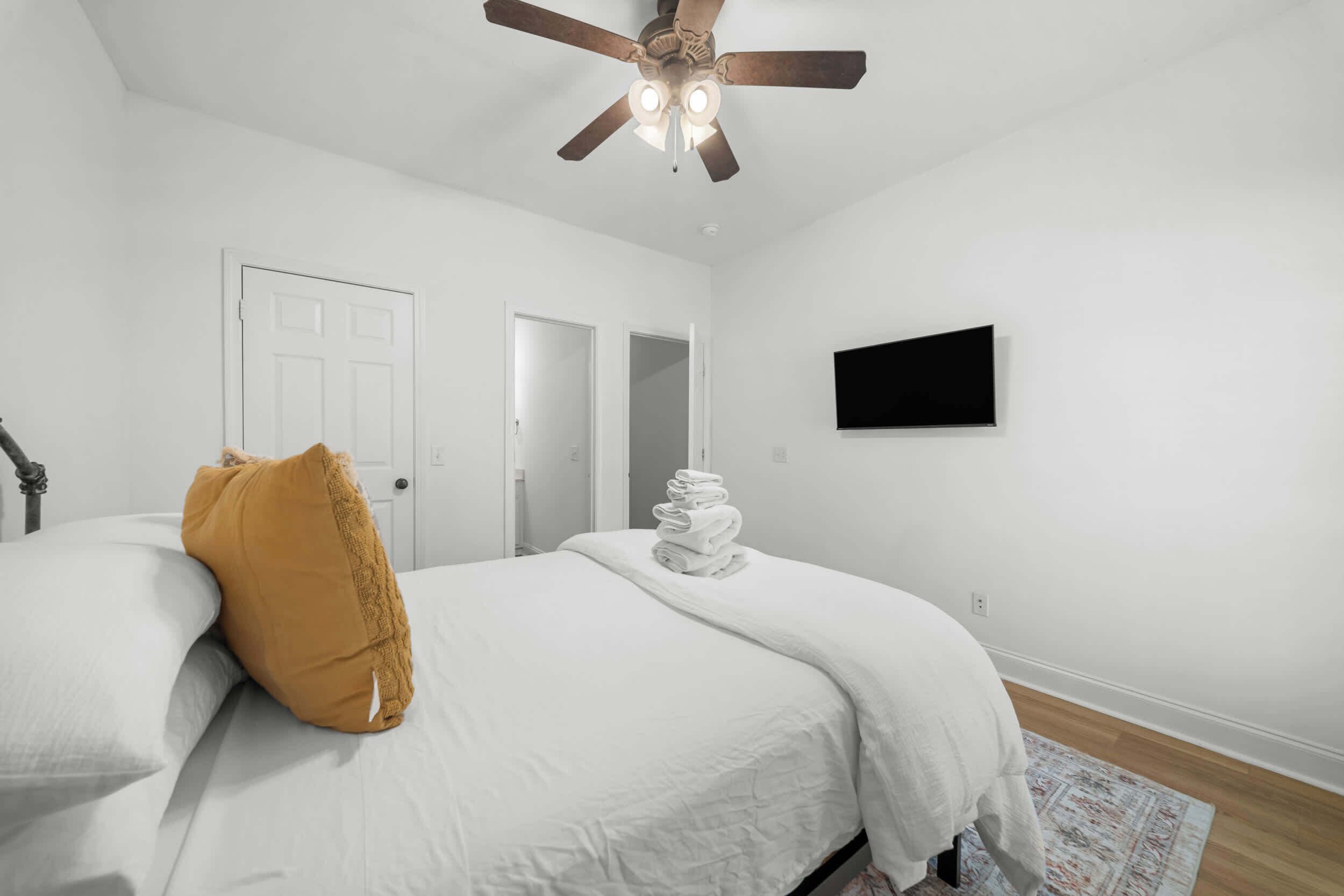 A bedroom with a bed, a ceiling fan, a white door, and a flat-screen TV on the wall.