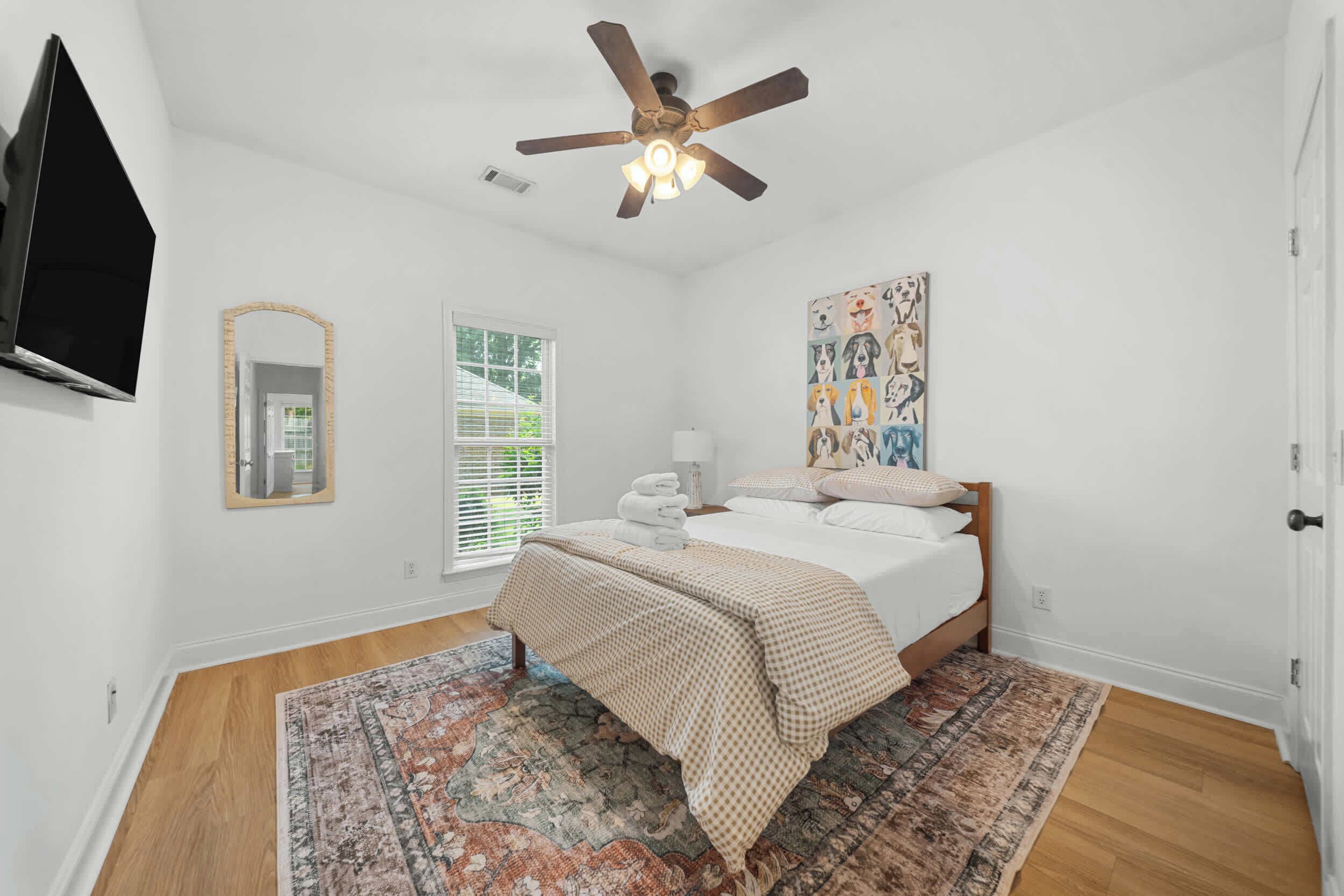 Une chambre à coucher avec un lit, un ventilateur de plafond, une fenêtre avec des stores, un tableau et un téléviseur fixé au mur.