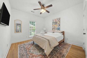 A bedroom with a bed, a ceiling fan, a window with blinds, a painting, and a television mounted on the wall.