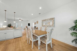 A spacious kitchen with white cabinets, a wooden dining table, and chairs. There is a large mirror on the wall and pendant lights above the table.