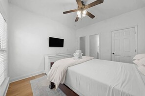 Un dormitorio con cama, televisión de pantalla plana montada en la pared, ventilador de techo y una puerta blanca.