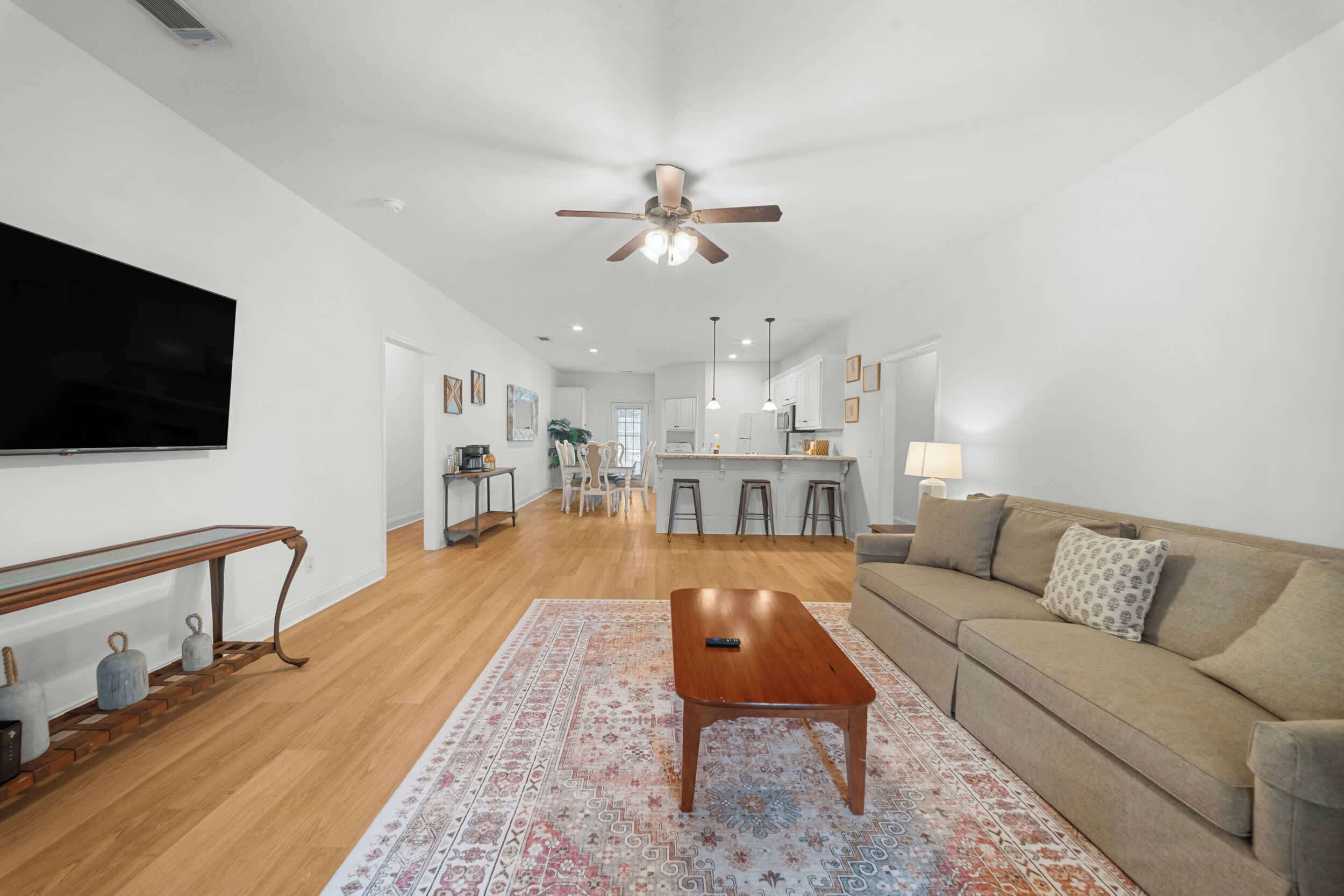 A spacious living room with a wooden coffee table, a beige sofa, and a large flat-screen TV mounted on the wall.