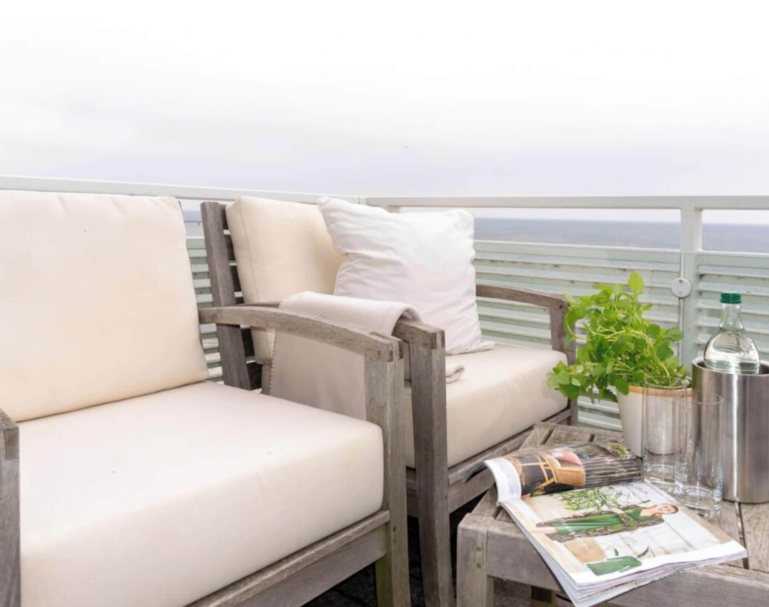 Un balcon avec une chaise en bois et un coussin, une plante en pot, un récipient en métal et un magazine.