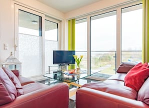 Ein modernes Wohnzimmer mit Ledersofas, einem Glastisch, einem Flachbildfernseher und Blick durch große Fenster.