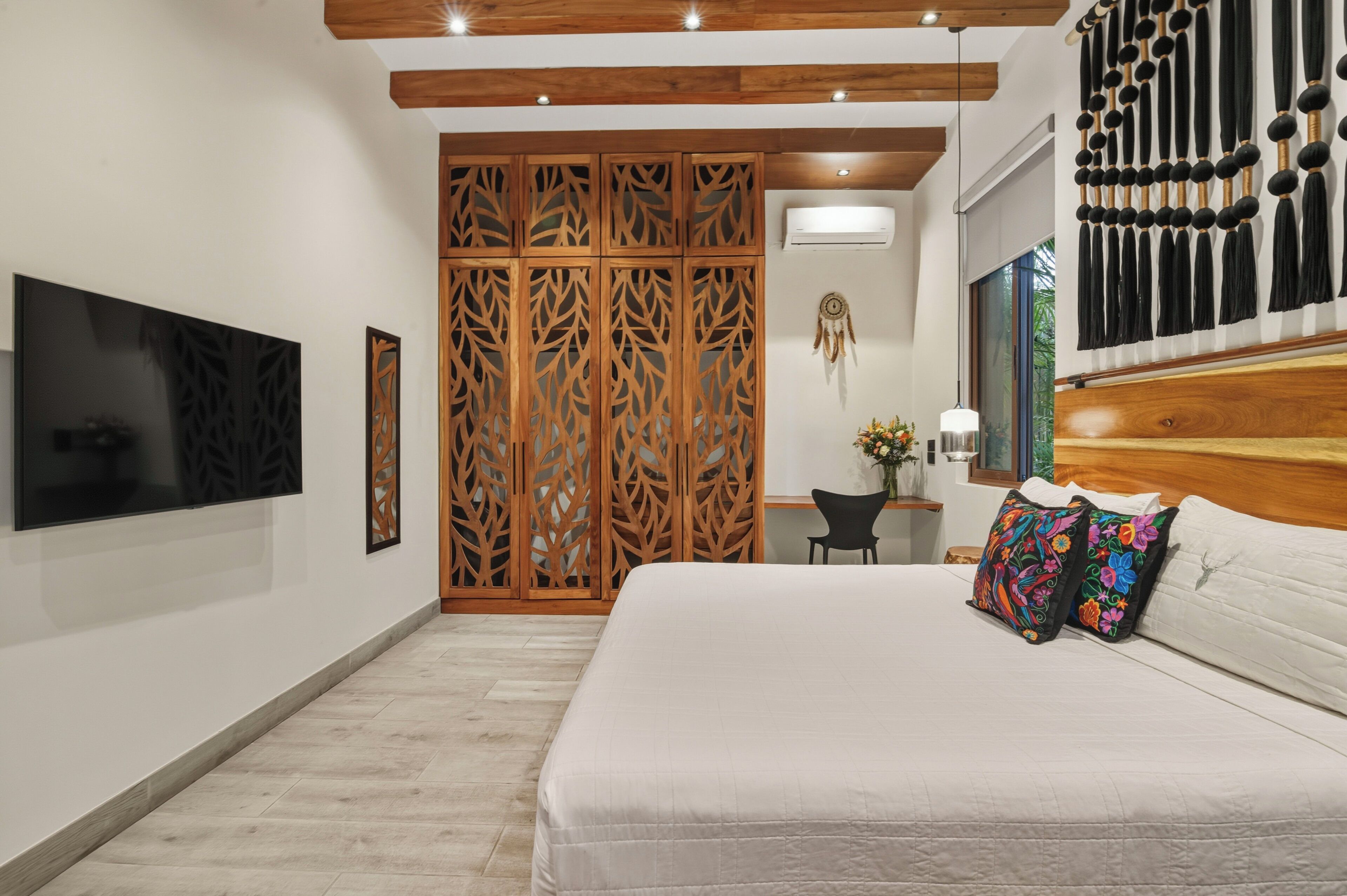 A bedroom with a bed, a TV mounted on the wall, a desk with a chair, and a decorative wall hanging.