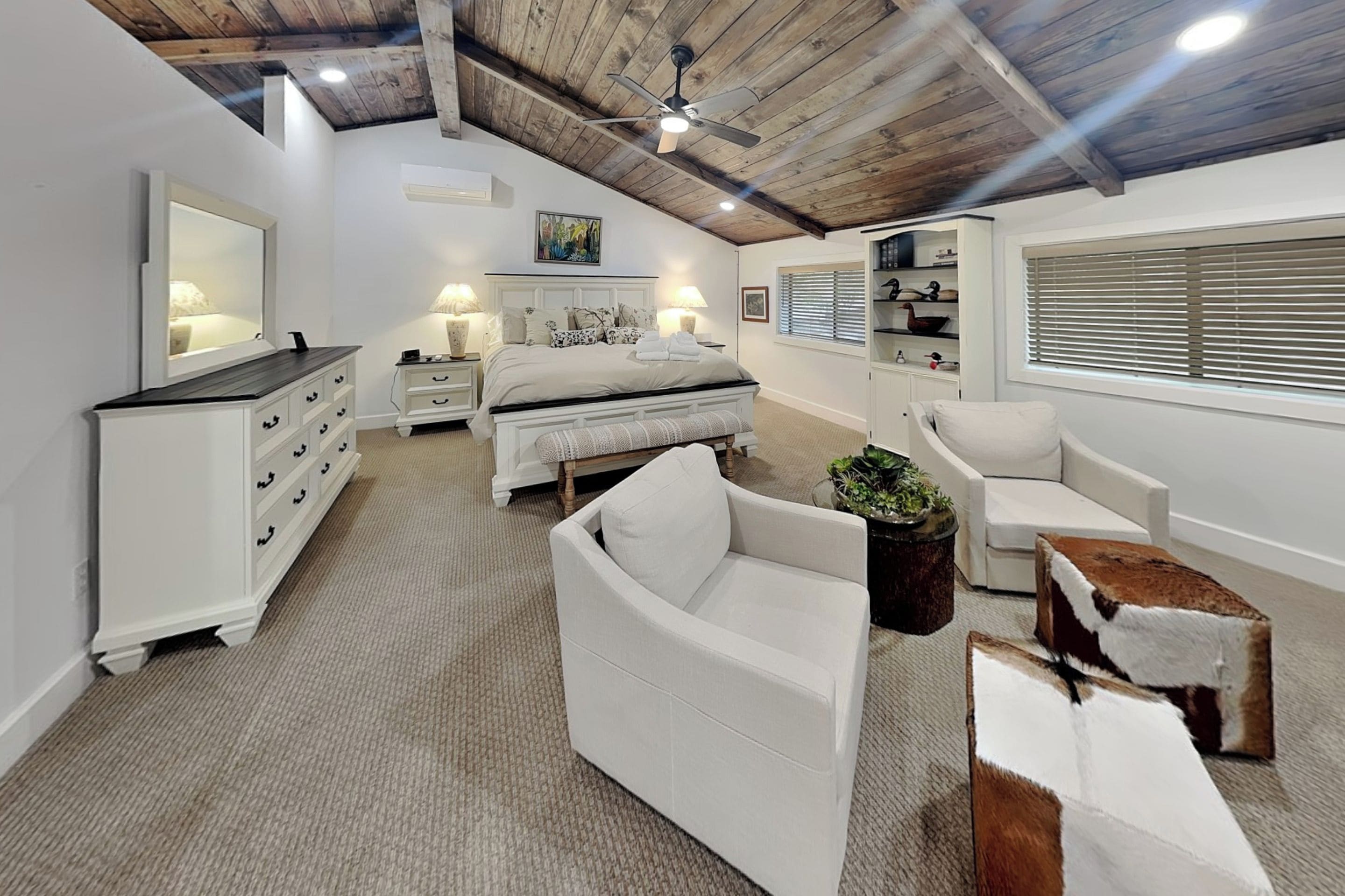 Un dormitorio con cama, mesitas de noche, cómoda, silla, una pequeña mesa y una planta en maceta.