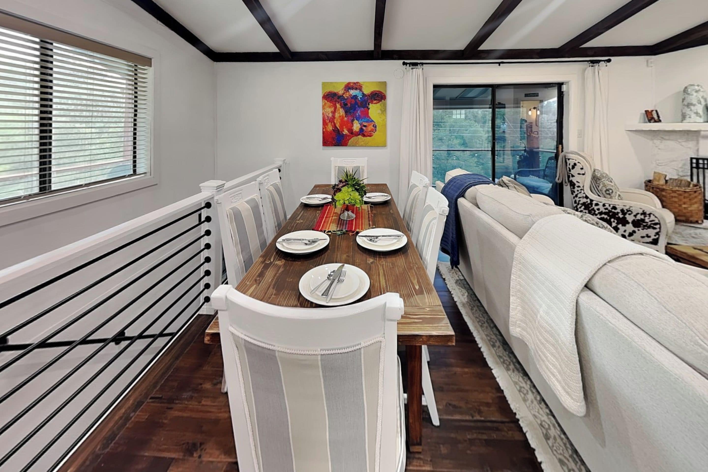 A dining area with a wooden table set for four, white chairs, a colorful painting on the wall, and a view through a glass door.