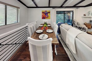 Uma área de refeições com mesa de madeira posta para quatro pessoas, cadeiras brancas, um quadro colorido na parede e vista através de uma porta de vidro.