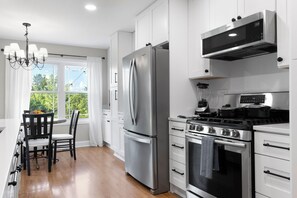 Cozinha moderna com eletrodomésticos de aço inoxidável, sala de jantar com lustre e vista para o verde lá fora.