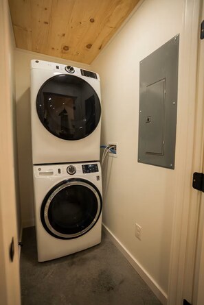 A laundry room with stacked washer and dryer units, a utility meter, and a door. - Casa Vetta | Modern Cabin with Fire Pit Near Davis (Davis)