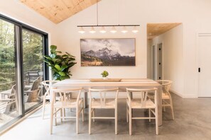 A dining area with a long table, multiple chairs, a large window, and a painting on the wall. - Casa Vetta | Modern Cabin with Fire Pit Near Davis (Davis)