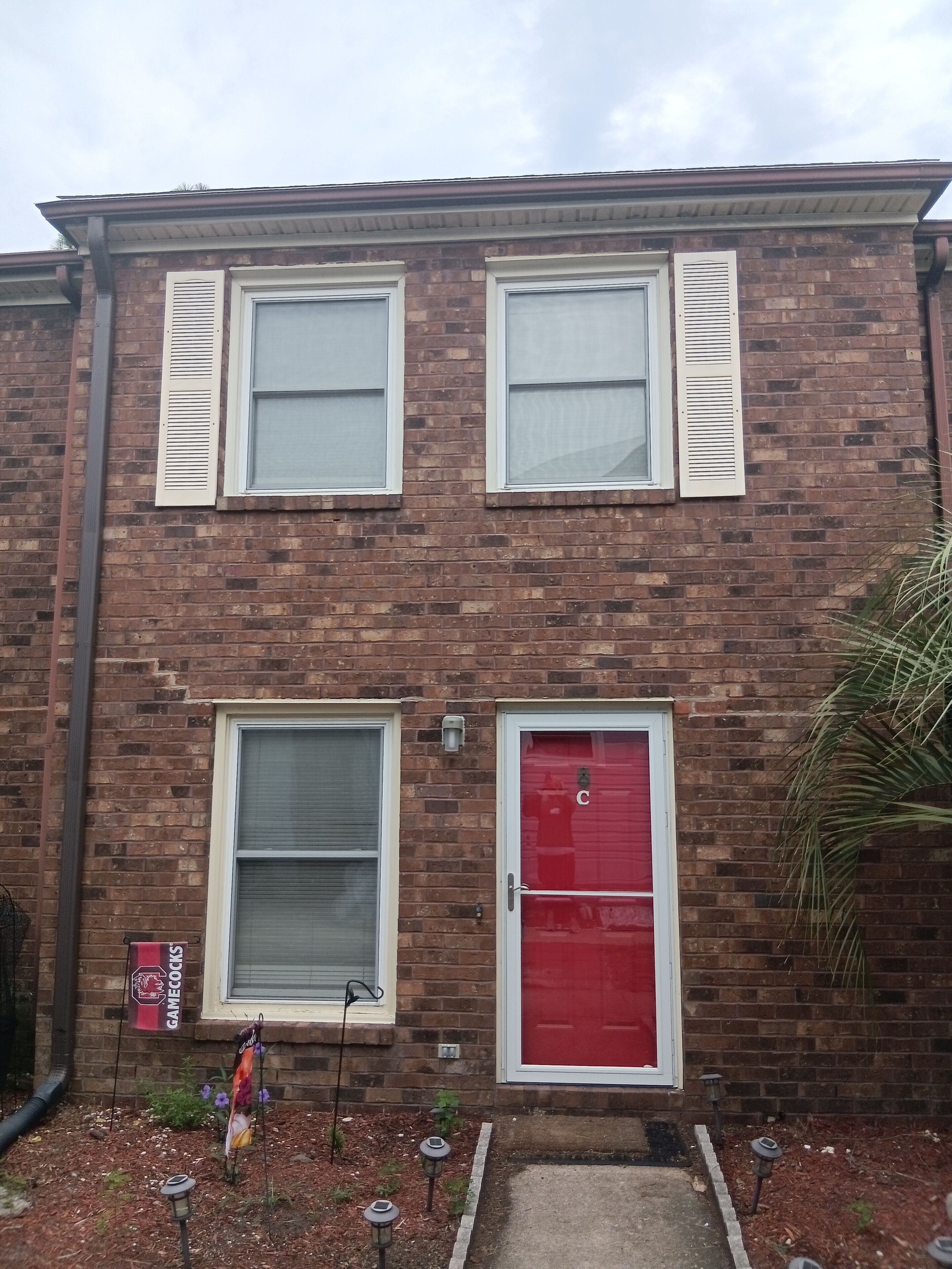 A two-story brick building with a red door, white shutters, and a small garden area with potted plants and outdoor lighting.