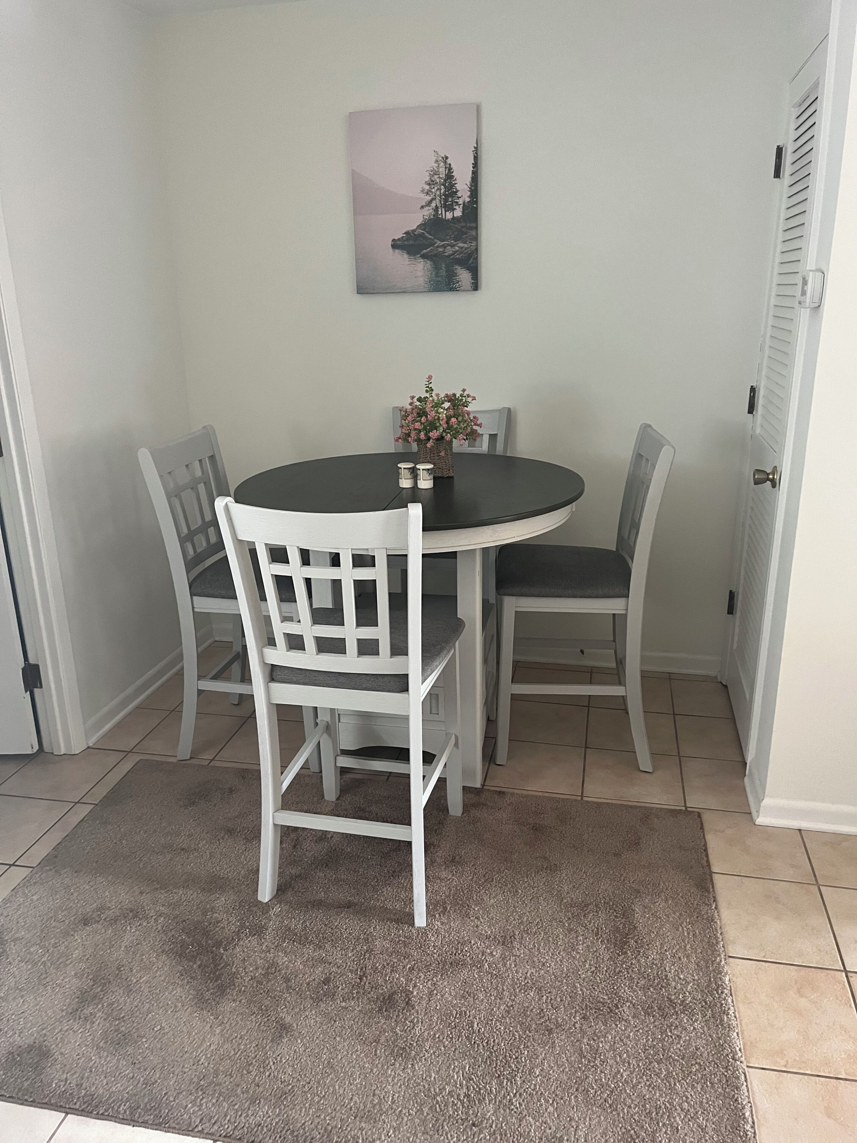 A dining area with a round table, four chairs, a small potted plant, and a painting on the wall.