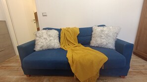 Un sofá azul con almohadones blancos y amarillos mullidos, ubicado en una habitación con piso de madera y una puerta blanca.