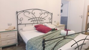 A bed with a metal headboard, white bedding, a red pillow, a green blanket, and a white towel. There is a bedside table with a book and a lamp. - Casa della Marmotta by Ca-sa Check (Aprica)