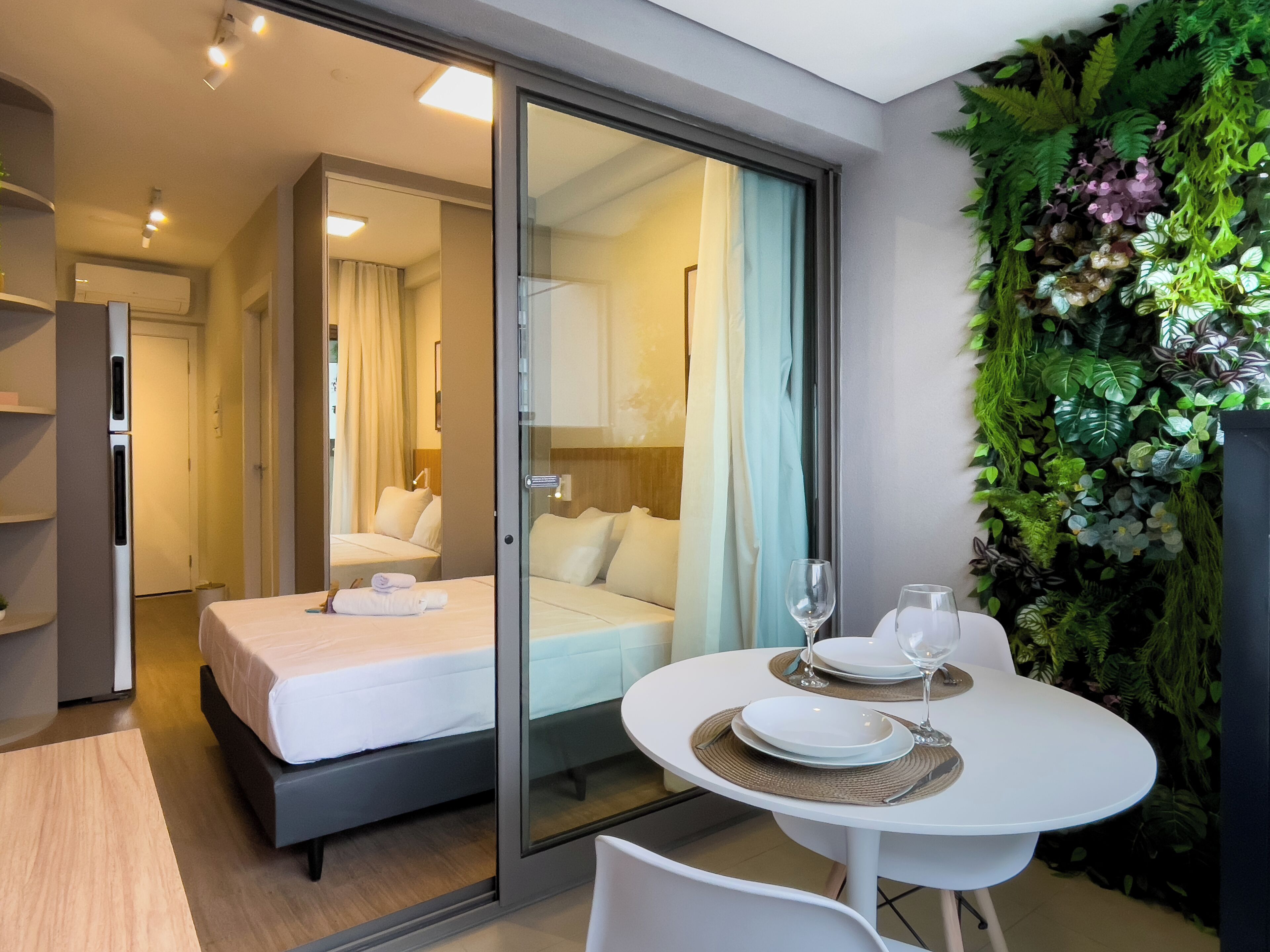 A modern hotel room with a glass door leading to a bedroom, a dining area with a table and chairs, and a green wall with plants.