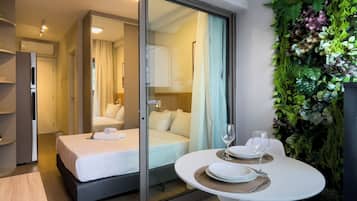 Una habitación de hotel moderna con una puerta de vidrio que da a un dormitorio, una zona de comedor con una mesa y sillas, y una pared verde con plantas.