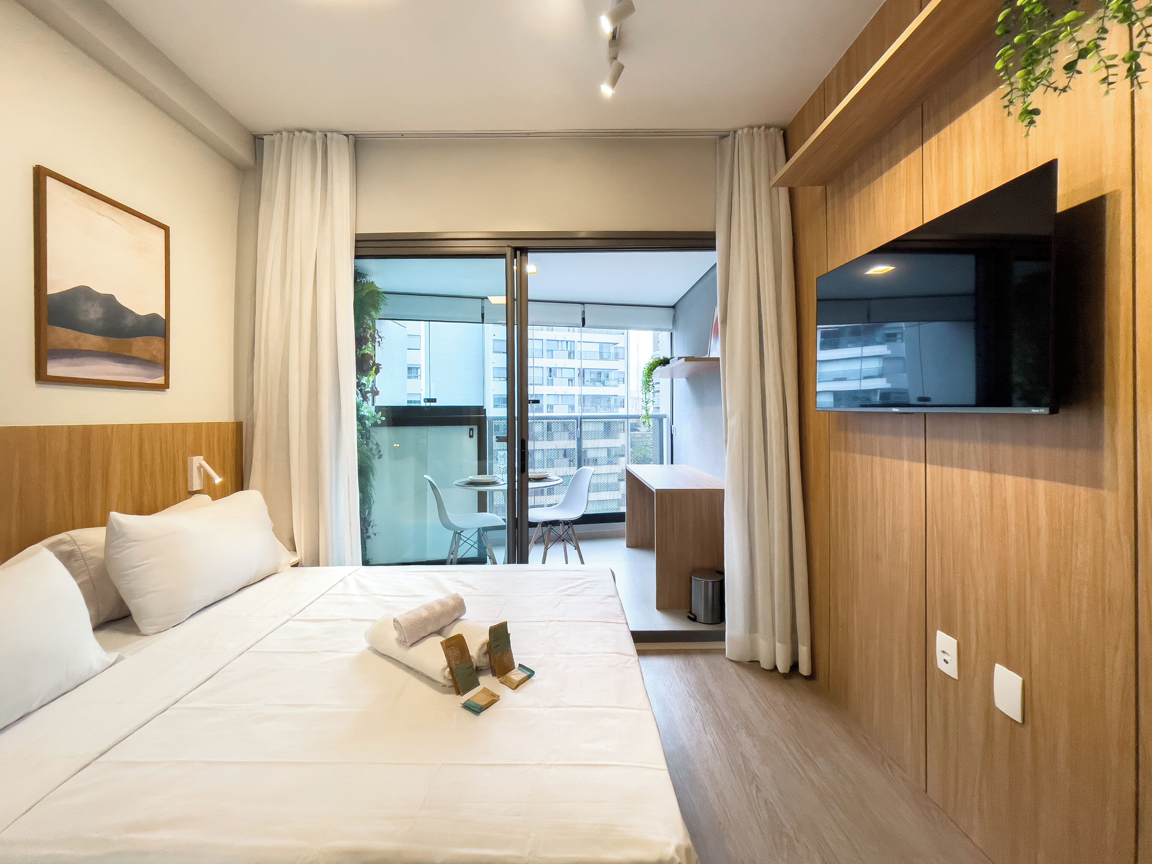 Habitación de hotel con cama, televisor montado en la pared, balcón con mesa y sillas, y un cuadro en la pared.