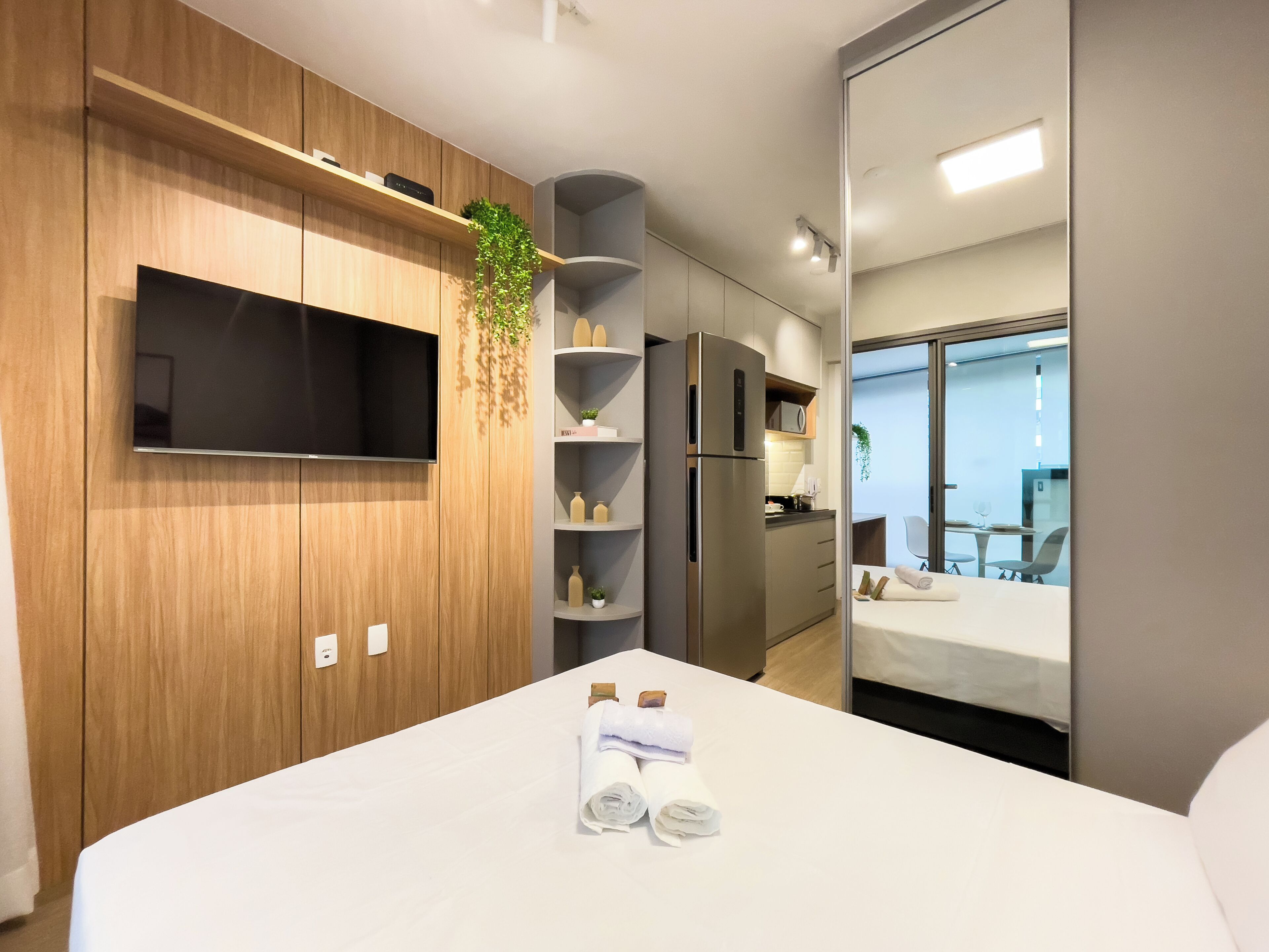 Habitación de hotel moderna con televisor de pantalla plana, paneles de madera, cocina integrada, balcón con asientos y una cama bien hecha.