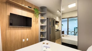 Habitación de hotel moderna con televisor de pantalla plana, paneles de madera, cocina integrada, balcón con asientos y una cama bien hecha.