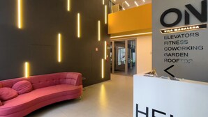 A modern interior with a red sofa, a reception desk, and a wall listing amenities like elevators, fitness, and coworking spaces. - ON Vila Madalena (São Paulo)