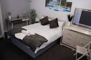 Ein ordentlich bezogenes Bett mit Kissen und einem gefalteten Handtuch, ein Nachttisch mit Lampe, ein kleiner Blumentopf und eine Kommode mit Schubladen.