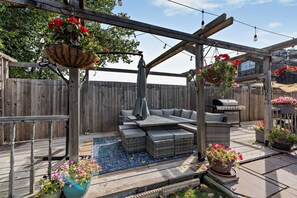 A wooden deck with a built-in seating area, a grill, hanging flower baskets, and a pergola structure. - Host & Stay | Thornbank (Chichester)